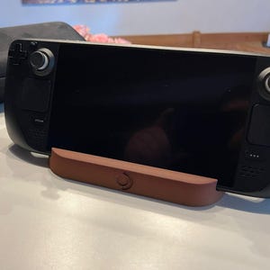 Könnte beinhalten: Schwarze Handheld-Spielkonsole mit großem Bildschirm auf einem braunen Ständer. Im Hintergrund befindet sich eine schwarze Tragetasche mit blauem Logo. Der Ständer ist etwa 15 cm lang.