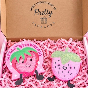 May include: A cardboard gift box filled with pink paper shreds containing two strawberry-shaped items. One is a flat card labelled Stella the Strawberry with a green top and pink face. The other is a textured pink strawberry bath bomb.