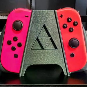 Op de afbeelding: Een op maat gemaakt gamingaccessoire met een metallic groene standaard, met een gestileerd "A"-ontwerp en een Triforce-symbool. Het houdt twee roze en rode gamecontrollers met zwarte knoppen, joysticks en directionele pads vast. De controllers zijn voor een gameconsole.