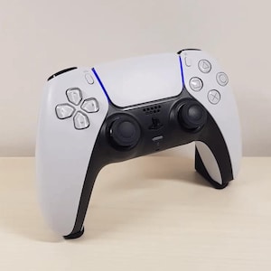 May include: A white and black PlayStation 5 DualSense wireless controller with a blue light bar. The controller has a touchpad, two analogue sticks, and a D-pad.