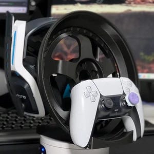 Puede incluir: Un soporte de auriculares para juegos blanco con un diseño de volante de carreras negro. El soporte tiene una luz azul y un mando blanco con un botón morado en el lado derecho.