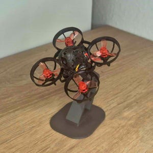 Può includere: Un piccolo drone nero e rosso con quattro eliche. Le eliche hanno pale rosse e sono circondate da telai neri. Il drone è montato su un supporto grigio, su una superficie di legno.
