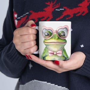 Puede incluir: Taza de cerámica blanca con una rana verde que lleva gafas de sol de corazón rosa y una pajarita rosa. La rana mira directamente al espectador.