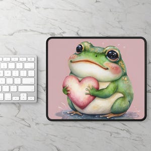 May include: A green frog with pink cheeks holds a pink heart in its hands. The frog is on a pink background with white speckles.