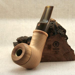 Puede incluir: Una pipa de fumar de madera con una boquilla de cuerno marrón. La pipa está apoyada en un trozo de madera con el logotipo de EPOS.