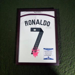Puede incluir: Camiseta enmarcada del Real Madrid con el nombre "RONALDO" y el número "7" en negro. La camiseta es blanca con detalles morados y una firma. Incluye un Certificado de Autenticidad Beckett y una pegatina "Compet Etsy".