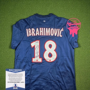 Pode incluir: Uma camisa de futebol azul marinho com o nome "IBRAHIMOVIC" e o número "18" em branco. A camisa tem uma assinatura e uma etiqueta "BECKETT CERTIFICATE OF AUTHENTICITY". A camisa está em uma superfície verde.