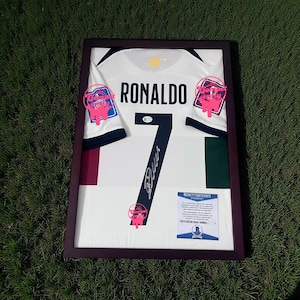 Może przedstawiać: Oprawiona biała koszulka piłkarska z nazwiskiem "RONALDO" i numerem "7" w kolorze czarnym. Koszulka posiada podpis i jest prezentowana w ciemnobrązowej ramie. Widoczny jest certyfikat autentyczności.