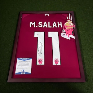 May include: Framed burgundy soccer jersey with the name "M. SALAH" and the number "11" in white. The jersey is signed and includes a Beckett Certificate. A pink and white ribbon with text is attached to the frame.