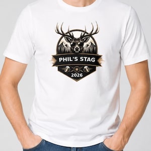 May include: A white short-sleeved t-shirt featuring a graphic of a stag wearing sunglasses in front of mountains. The design includes a black shield emblem with the text PHIL'S STAG, two beer mugs, a star, and the year 2026 printed in white.