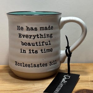 May include: Cream-colored ceramic mug with a teal rim and a tan base. The mug features the text "He has made Everything beautiful in its time, Ecclesiastes 3:11." A black tag is attached to the handle.