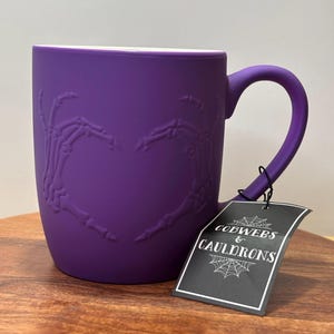 Pode incluir: Uma caneca roxa com um design de mãos de esqueleto formando um coração. A caneca tem uma alça e uma etiqueta que diz "COBWEBS & CAULDRONS". A caneca está em uma superfície de madeira.