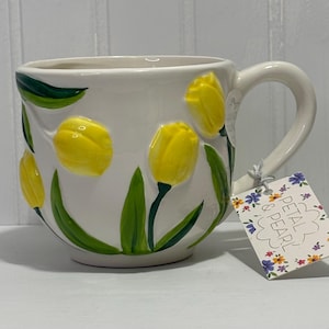Puede incluir: Taza de cerámica blanca con flores de tulipán amarillas en relieve y hojas verdes. La taza tiene un asa curva y una etiqueta decorativa que dice "Petal & Pearl". La taza está diseñada con un tema floral.