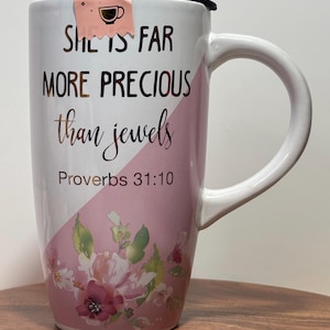 May include: White ceramic mug with a pink and white floral design. The mug has a black lid and a handle. The text on the mug reads "She is far more precious than jewels Proverbs 31:10".