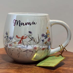 Könnte beinhalten: Weiße Keramiktasse mit Blumenmuster und dem Wort "Mamá" in rosa Schrift. Die Tasse hat eine abgerundete Form und einen goldenen Henkel.