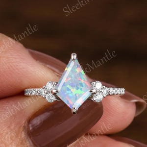 May include: A silver ring featuring a kite-shaped opal center stone with iridescent colors. The ring is accented with small, round, clear gemstones on the band and on either side of the opal. The ring is held up by a person's hand.