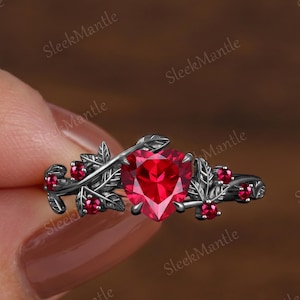 May include: A silver-toned ring featuring a heart-shaped, vibrant red gemstone as the centerpiece. The ring is adorned with leaf and berry details, creating a nature-inspired design. Small red gemstones accent the leaves and branches.