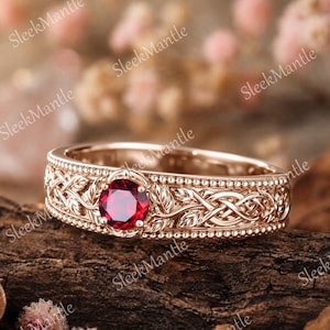 Puede incluir: Anillo de oro rosa con una piedra preciosa roja oscura redonda en el centro. La banda presenta intrincados diseños de nudos celtas y hojas, con un borde de cuentas. El anillo se muestra sobre un trozo de madera.
