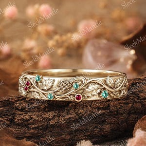 May include: A gold-colored ring with an intricate design featuring leaves, berries, and gemstones. The ring has a band with detailed carvings and is adorned with red and teal-colored gemstones. The ring is displayed on a piece of wood.