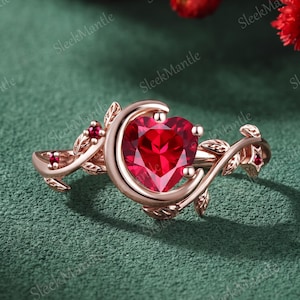 May include: A rose gold ring featuring a central heart-shaped red gemstone cradled by a crescent moon shape. The band is designed with intricate vine and leaf motifs, accented with small round red stones embedded along the metal branches.