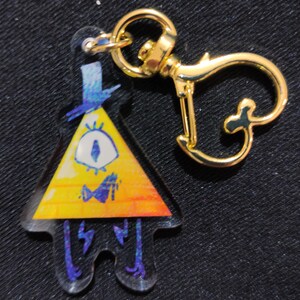 Può includere: Un portachiavi dorato con un ciondolo giallo a forma di triangolo che presenta un personaggio dei cartoni animati con un solo occhio, un papillon e un cappello a cilindro blu. Il personaggio è Bill Cipher della serie animata Gravity Falls.