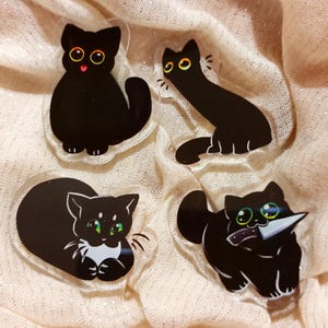 以下が含まれることがあります： 異なる表情の黒い猫のアクリルチャームが4つ。1匹は尻尾を脚に巻き付けて座り、もう1匹は尻尾をまっすぐ伸ばして座り、もう1匹は顎の下に足を折り畳んで横になり、最後の1匹は口にナイフを持って立っている。