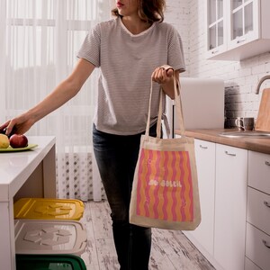 Un tote bag coloré de vagues roses et oranges, une inscription "au soleil" dessinée en blanc en arrondi au milieu et autour des petits motifs dessinés en blanc, un soleil, des lunettes de soleil, un appareil photo, un cornet de glace, une étoile de mer et une paire de tongs. Une femme tient le sac d'une main devant un plan de travail dans une cuisine, de l’autre main elle pose des des fruits dans une assiette.