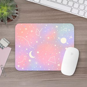 May include: A rectangular mousepad with a pastel pink, purple, and blue gradient background. White constellations, stars, a crescent moon, and a planet with rings are scattered across the surface. A white computer mouse rests on the right side.