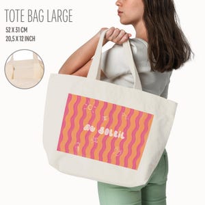 Un tote bag coloré de vagues roses et oranges, une inscription "au soleil" dessinée en blanc en arrondi au milieu et autour des petits motifs dessinés en blanc, un soleil, des lunettes de soleil, un appareil photo, un cornet de glace, une étoile de mer et une paire de tongs. Le sac est le modèle large, il est porté par une femme en tee-shirt blanc. des infos sur la dimension et une petite photo de la poche intérieur sur le côté. Le fond est neutre.