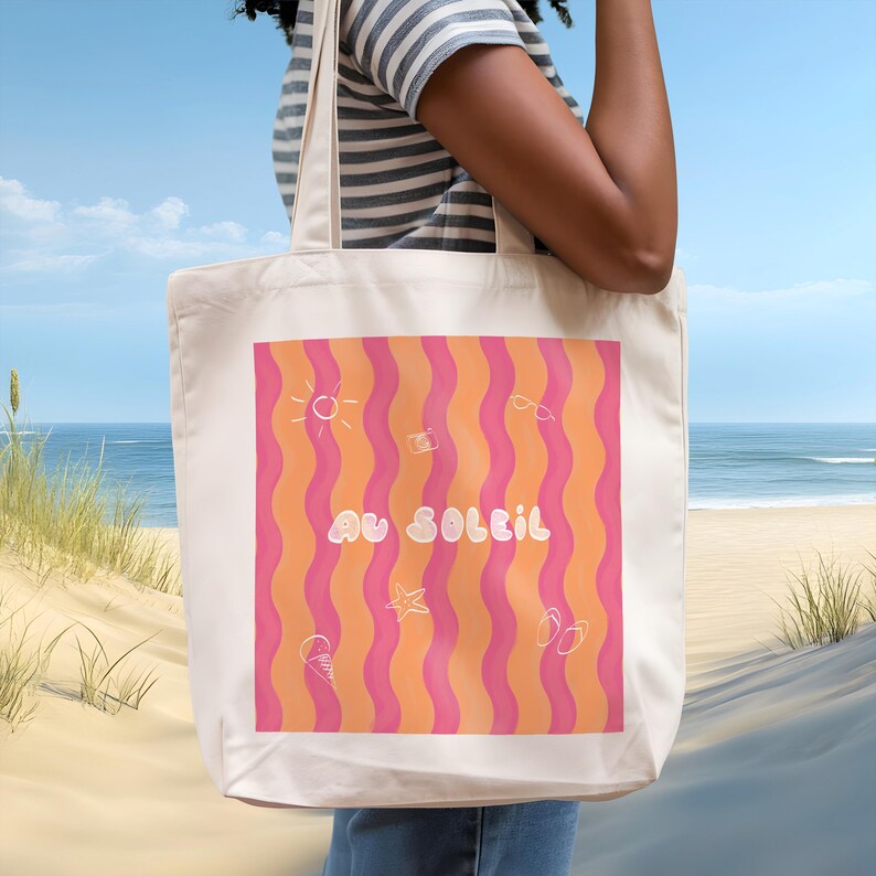 Un tote bag coloré de vagues roses et oranges, une inscription "au soleil" dessinée en blanc en arrondi au milieu et autour des petits motifs dessinés en blanc, un soleil, des lunettes de soleil, un appareil photo, un cornet de glace, une étoile de mer et une paire de tongs. Une femme porte le sac devant une plage, sable fin, mer calme et ciel bleu en arrière plan.