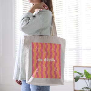 Un tote bag coloré de vagues roses et oranges, une inscription "au soleil" dessinée en blanc en arrondi au milieu et autour des petits motifs dessinés en blanc, un soleil, des lunettes de soleil, un appareil photo, un cornet de glace, une étoile de mer et une paire de tongs. Une femme en chemise à rayures bleues et blanches porte le sac, elle est dans une pièce lumineuse devant une fenêtre.