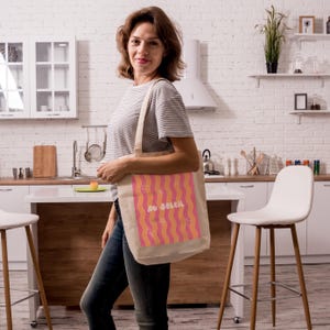 Un tote bag coloré de vagues roses et oranges, une inscription "au soleil" dessinée en blanc en arrondi au milieu et autour des petits motifs dessinés en blanc, un soleil, des lunettes de soleil, un appareil photo, un cornet de glace, une étoile de mer et une paire de tongs. Une femme qui sourit porte le sac dans une cuisine chaleureuse et moderne.