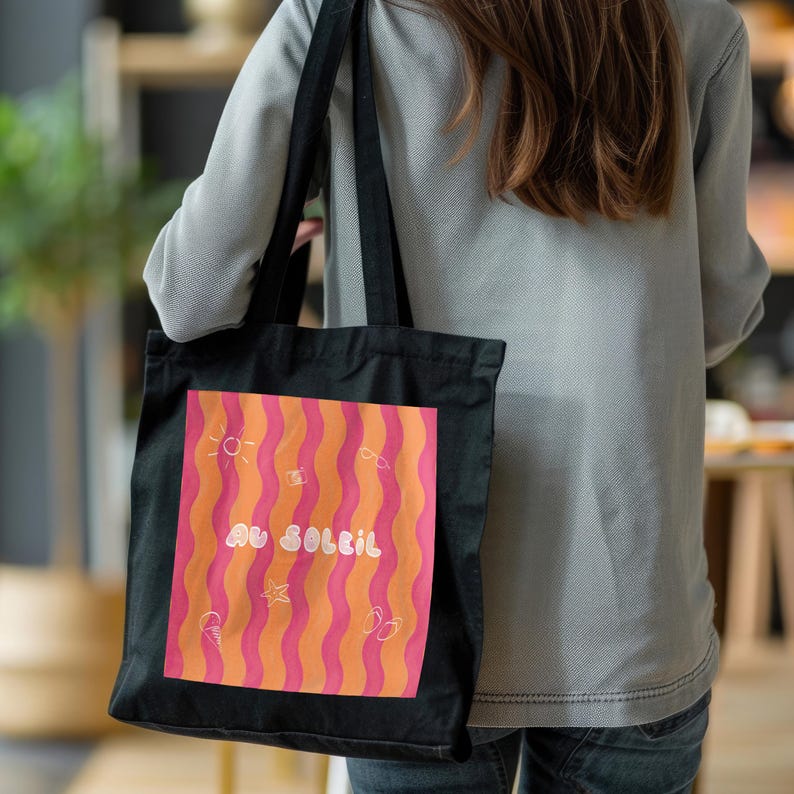 Un tote bag coloré de vagues roses et oranges, une inscription "au soleil" dessinée en blanc en arrondi au milieu et autour des petits motifs dessinés en blanc, un soleil, des lunettes de soleil, un appareil photo, un cornet de glace, une étoile de mer et une paire de tongs.  
Une femme en chemise bleu ciel et aux cheveux long porte le tote bag noir à l’épaule. Elle est dans un restaurant à la lumière chaleureuse.