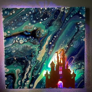 May include: Square canvas art with an abstract fluid design in shades of teal, blue, and gold. A silhouette of a castle is cut out and backlit with warm and cool colored lights, creating a magical effect. The artwork is illuminated with a purple border.