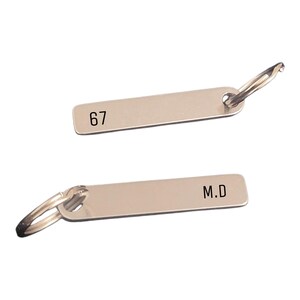 Peut inclure: Deux porte-clés argentés avec des étiquettes rectangulaires. L'une porte le numéro "67", l'autre les initiales "M.D.". Chaque porte-clés est muni d'un fermoir métallique.