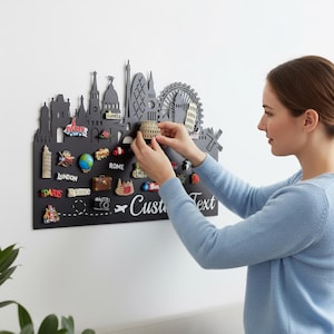 May include: A black wall-mounted travel-themed display board with a city skyline silhouette and various miniature souvenir magnets. The board has the text "Custom Text" and a woman is placing a magnet on the board.