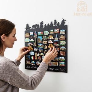 May include: A black magnetic board with a silhouette of a group of figures at the top, holding a collection of colorful souvenir magnets. The magnets depict various landmarks and buildings. The text "Not all those who wander are lost" is at the bottom.