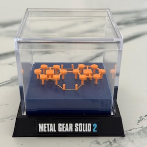 May include: An orange geometric sculpture inside a clear plastic display case. The sculpture sits on a blue base within the case, which is mounted on a black base with the text "METAL GEAR SOLID 2".