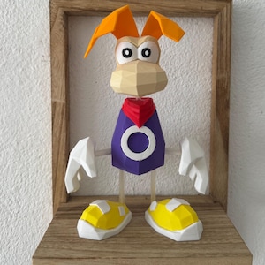 May include: A low-poly 3D printed figurine of the character Rayman, featuring a tan head with orange hair, a purple torso with a white circular emblem, a red neckerchief, white gloves, and yellow shoes with white accents, standing on a wooden shelf.