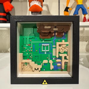 May include: A framed 3D map of a video game world, featuring green, blue, and tan terrain. The map is encased in a black frame with a yellow triangle emblem at the bottom. The artwork is a detailed, layered representation of a classic game.