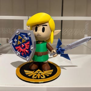 Könnte beinhalten: Figur eines Charakters aus der Videospielreihe The Legend of Zelda. Die Figur hat eine gelbe Kappe, eine grüne Tunika und braune Stiefel. Sie hält einen Schild und ein Schwert. Der Sockel ist schwarz und goldfarben mit einem Wappen.