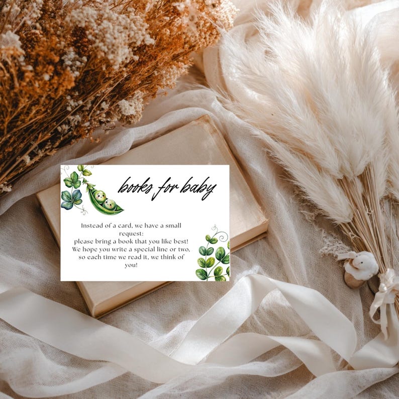 May include: A white card with the text "books for baby" and a request to bring a book. The card features a watercolor illustration of a pea pod with two peas and green vines. The card is on top of a book.