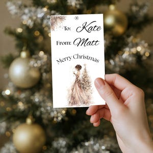 May include: A white Christmas gift tag with the text "To: Kate, From: Matt, Merry Christmas." The tag features a watercolour illustration of a woman in a gown next to a Christmas tree. The tag is held in front of a Christmas tree.