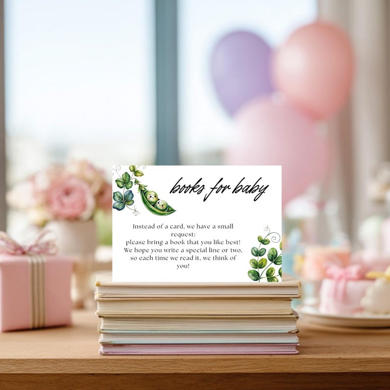 May include: A white card with the text "books for baby" and a request for guests to bring a book. The card features a watercolor illustration of peas in a pod and green vines. The card is placed on a stack of books.