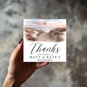 May include: A square wedding favour card with a beach scene showing a wooden walkway to the sea. The card says "Thanks for coming to Matt & Kate's Wedding" in elegant script.