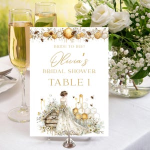 May include: A white table card with gold text that reads "Bride to Bee! Olivia's Bridal Shower Table 1." The card features a watercolor illustration of a bride, beehive, and honeycomb design. Two champagne flutes and a bouquet of white flowers are in the background.