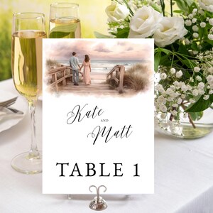 May include: A white table card with the names "Kate and Matt" and "TABLE 1" in black script. The card features a watercolour painting of a couple walking on a beach boardwalk. A silver card holder and champagne glasses are in the foreground.