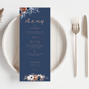 May include: A navy blue wedding menu card with floral accents in shades of blue, brown, and white. The menu includes the word "Menu" in a copper-coloured script, along with the entree, main course, and dessert options.