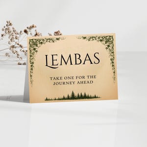 May include: A rectangular card with the word "LEMBAS" in a bold, black font. The card has a vintage parchment paper appearance, with green decorative borders and the text "TAKE ONE FOR THE JOURNEY AHEAD". A row of green trees is at the bottom.
