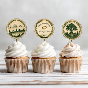 May include: Three cupcakes with white frosting and themed toppers. The toppers feature circular designs with illustrations of mountains, trees, and the text "ONE BIRTHDAY TO RULE THEM ALL". The cupcakes sit on a light-colored wooden surface.